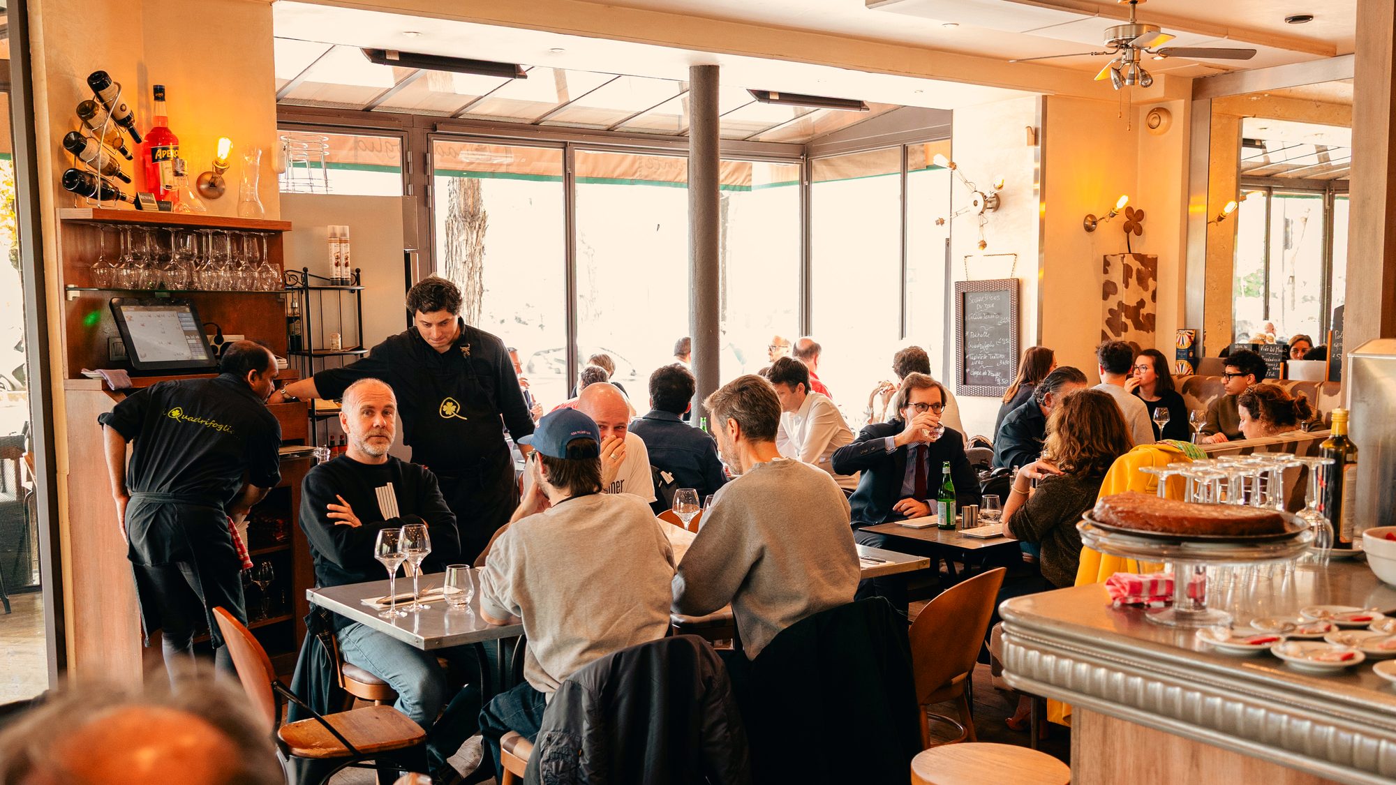 Salle du restaurant en plein service, comptoir en bois et grandes baies vitrées.